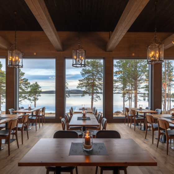 Viihtyisä ravintola, jossa on puupöytiä ja -tuoleja, sarvikruunut ja suuret ikkunat, joista avautuu näkymä järvelle ja mäntyihin auringonlaskun aikaan. Lämmin valaistus luo kutsuvan ilmapiirin.