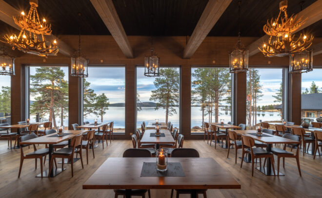 Viihtyisä ravintola, jossa on puupöytiä ja -tuoleja, sarvikruunut ja suuret ikkunat, joista avautuu näkymä järvelle ja mäntyihin auringonlaskun aikaan. Lämmin valaistus luo kutsuvan ilmapiirin.