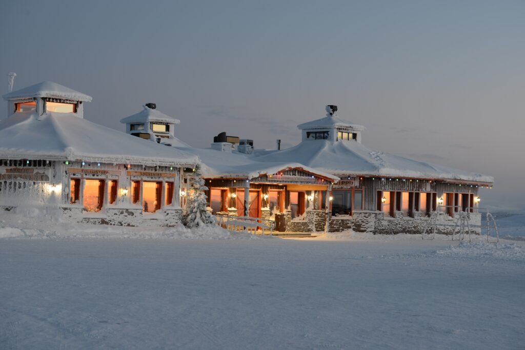 Suuri, lämpimästi valaistu, lumen ja jääpuikkojen peittämä puinen mökki seisoo lumisessa maisemassa iltahämärässä, ja taustalla on pehmeän sininen taivas.