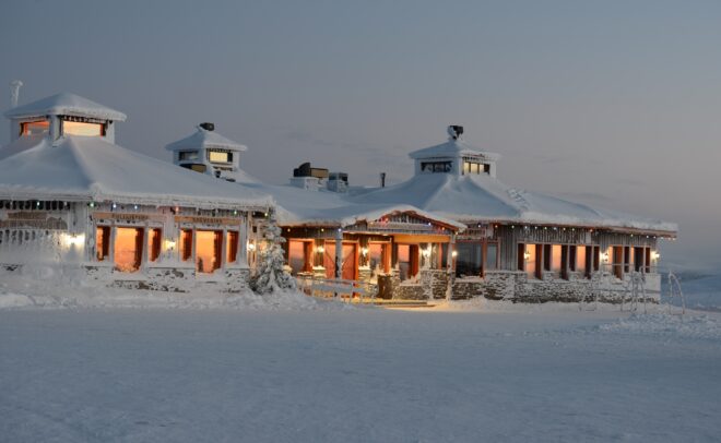 Suuri, lämpimästi valaistu, lumen ja jääpuikkojen peittämä puinen mökki seisoo lumisessa maisemassa iltahämärässä, ja taustalla on pehmeän sininen taivas.