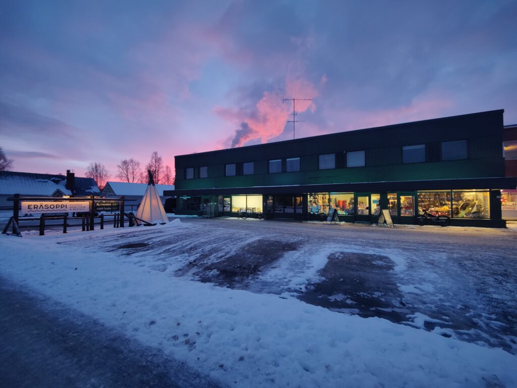 Lumipeitteinen kaupunkimaisema auringonlaskun aikaan, jossa näkyy kaksikerroksinen rakennus, jonka sisällä loistaa kirkkaita valoja, pieni tiipiimäinen rakenne sekä taivaalla värikkäät pilvet.