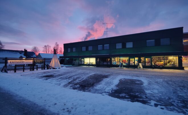 Lumipeitteinen kaupunkimaisema auringonlaskun aikaan, jossa näkyy kaksikerroksinen rakennus, jonka sisällä loistaa kirkkaita valoja, pieni tiipiimäinen rakenne sekä taivaalla värikkäät pilvet.