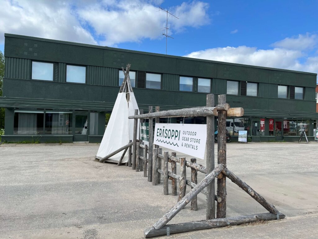 Vihreä kaksikerroksinen rakennus, jonka etupuolella on puukehyksessä kyltti, jossa lukee ”Erisoppi Outdoor Gear Store & Rentals”, ja sen vieressä valkoinen kankaasta tehty tiipi. Taivas on osittain pilvinen.
