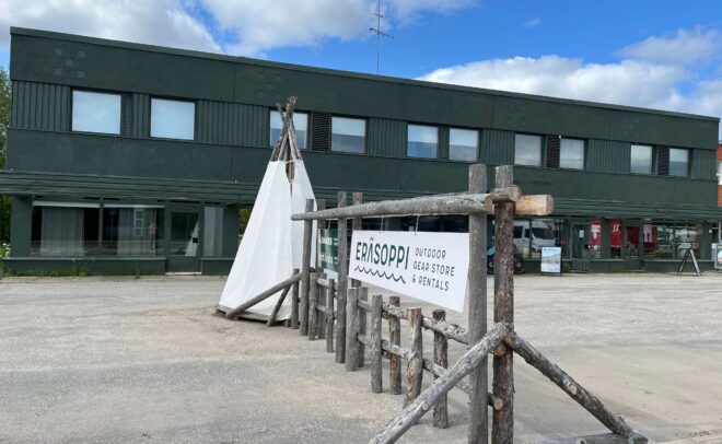 Vihreä kaksikerroksinen rakennus, jonka etupuolella on puukehyksessä kyltti, jossa lukee ”Erisoppi Outdoor Gear Store & Rentals”, ja sen vieressä valkoinen kankaasta tehty tiipi. Taivas on osittain pilvinen.