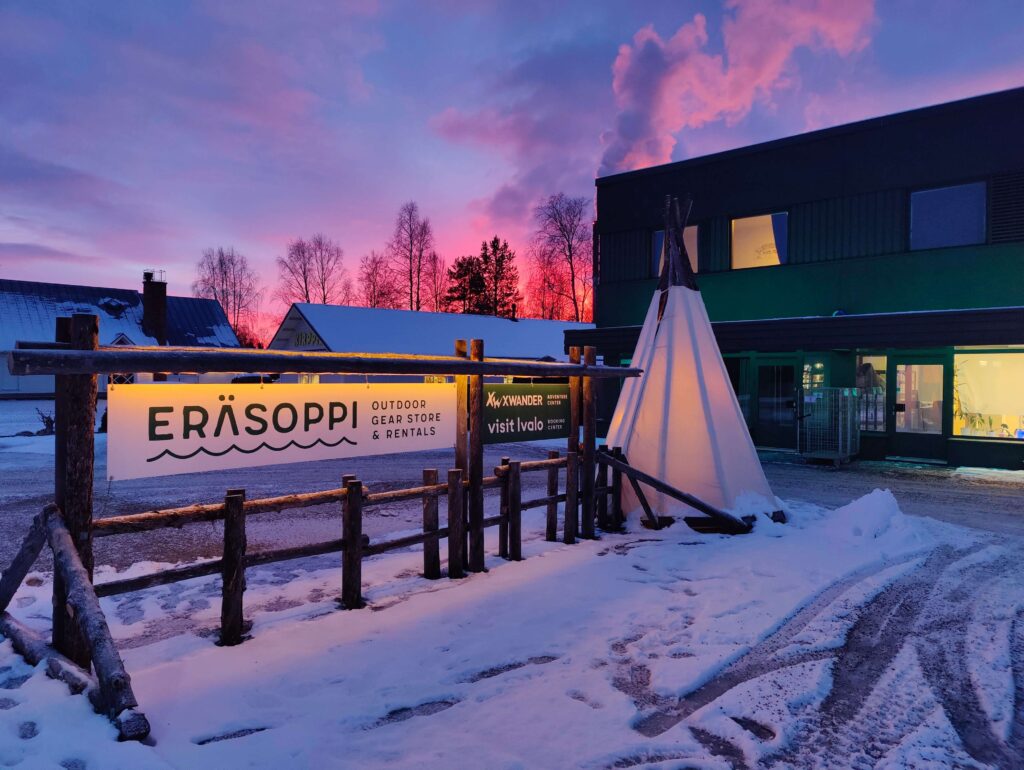 Eräsoppi-ulkoiluvälinekauppa, jossa on puinen kyltti ja valkoinen tipi auringonlaskun aikaan lumisessa maisemassa; taustalla näkyy rakennus ja puita värikkään vaaleanpunaisen ja violetin taivaan alla.