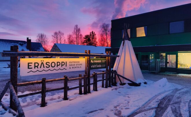 Eräsoppi-ulkoiluvälinekauppa, jossa on puinen kyltti ja valkoinen tipi auringonlaskun aikaan lumisessa maisemassa; taustalla näkyy rakennus ja puita värikkään vaaleanpunaisen ja violetin taivaan alla.