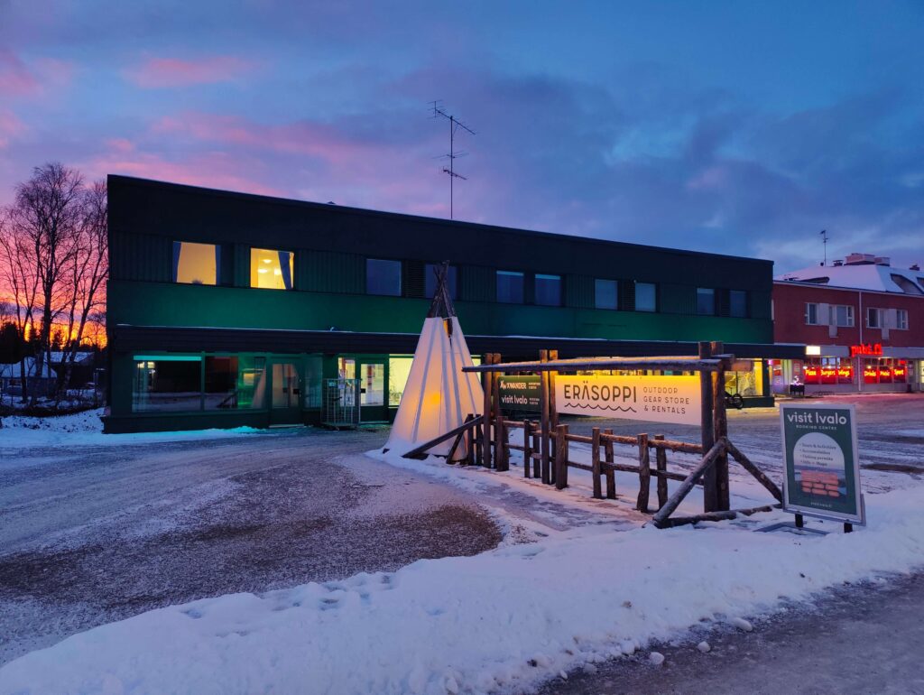 Hämärässä lumiin kätkeytyy moderni, kaksikerroksinen ympäristöystävällinen rakennus, jonka ikkunat loistavat valaistuina. Sen edessä näkyy valaistu tiipi-rakennelma sekä Eräsoppi-ulkoilukaupan puinen kyltti. Vaaleanpunainen ja sininen taivas luo rauhallisen tunnelman.
