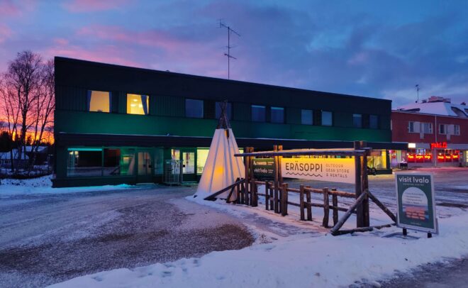 Hämärässä lumiin kätkeytyy moderni, kaksikerroksinen ympäristöystävällinen rakennus, jonka ikkunat loistavat valaistuina. Sen edessä näkyy valaistu tiipi-rakennelma sekä Eräsoppi-ulkoilukaupan puinen kyltti. Vaaleanpunainen ja sininen taivas luo rauhallisen tunnelman.