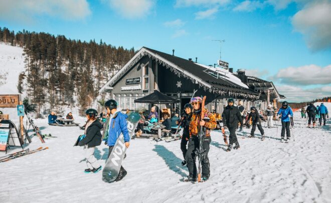 Talvivaatteisiin pukeutuneet hiihtäjät ja lumilautailijat ovat kokoontuneet lumisen vuoristomajan eteen, jossa ihmiset istuvat pöydän ääressä. Taustalla näkyy mäntyjä ja sinistä taivasta, mikä luo eloisan ja kauniin talvimaiseman.