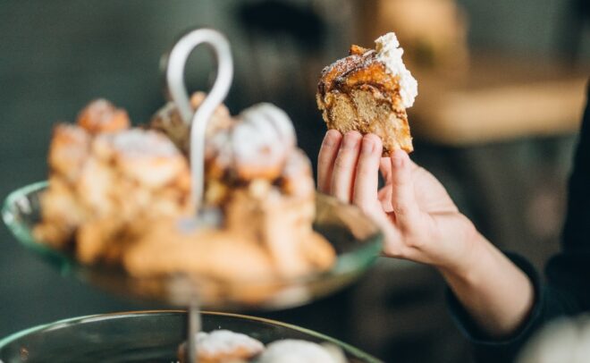 Käsi, joka pitää kädessään palan kuorrutettua kanelirullaa, lasitelineen yläpuolella, joka on täynnä muita leivonnaisia ja kanelirullia pehmeästi sumennetussa taustassa.