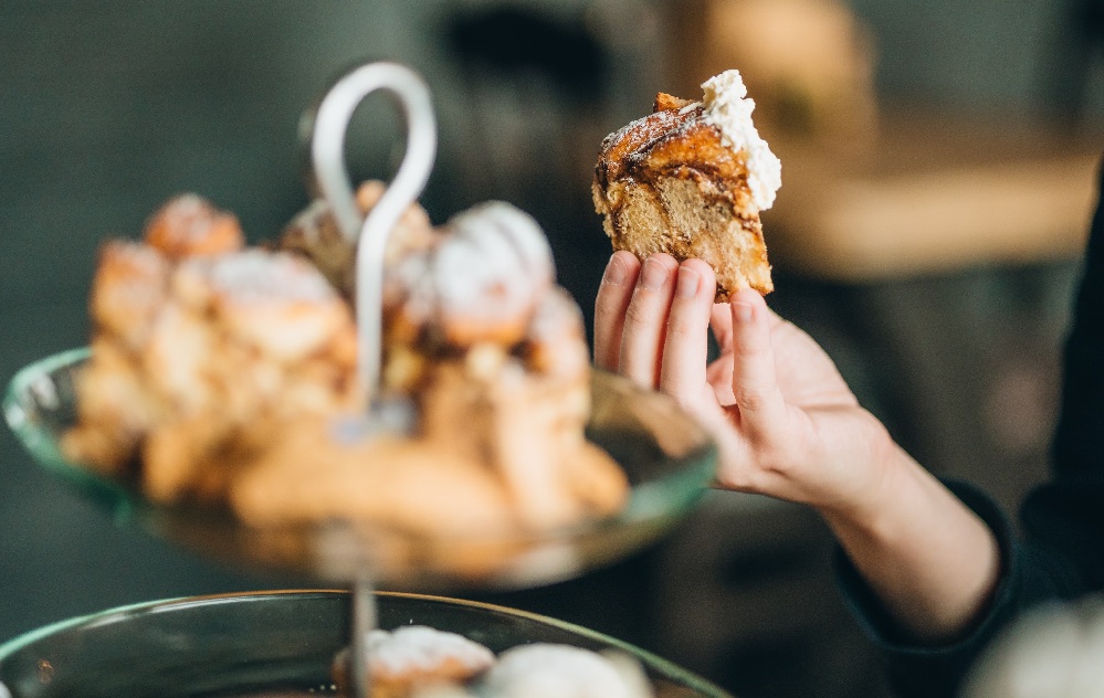 Käsi, joka pitää kädessään palan kuorrutettua kanelirullaa, lasitelineen yläpuolella, joka on täynnä muita leivonnaisia ja kanelirullia pehmeästi sumennetussa taustassa.