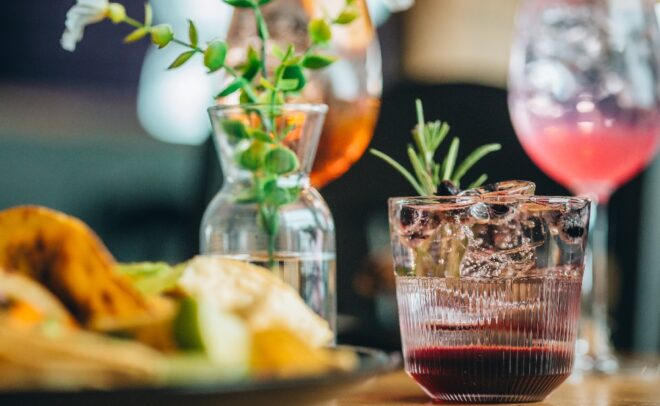 Lähikuva lasillisesta jääkylmää punaista cocktailia, joka on koristeltu rosmariinilla; taustalla näkyy epäselviä kukkia, toinen juoma ja ruokalautanen.