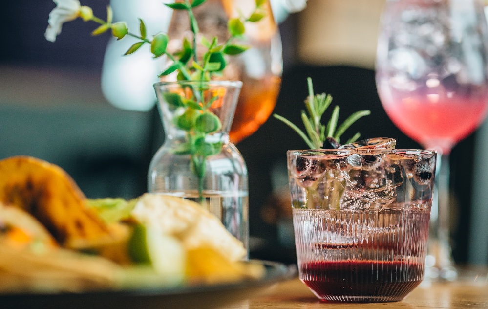 Lähikuva lasillisesta jääkylmää punaista cocktailia, joka on koristeltu rosmariinilla; taustalla näkyy epäselviä kukkia, toinen juoma ja ruokalautanen.