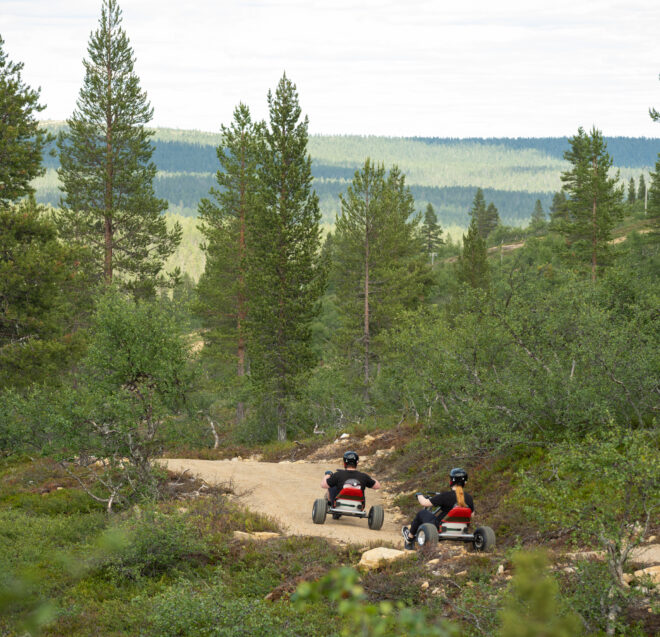 Kaksi ihmistä ajaa polkuautoilla mutkittelevaa hiekkatietä pitkin rehevän vihreän metsän läpi, puiden ja kukkuloiden ympäröimänä pilvisen taivaan alla.