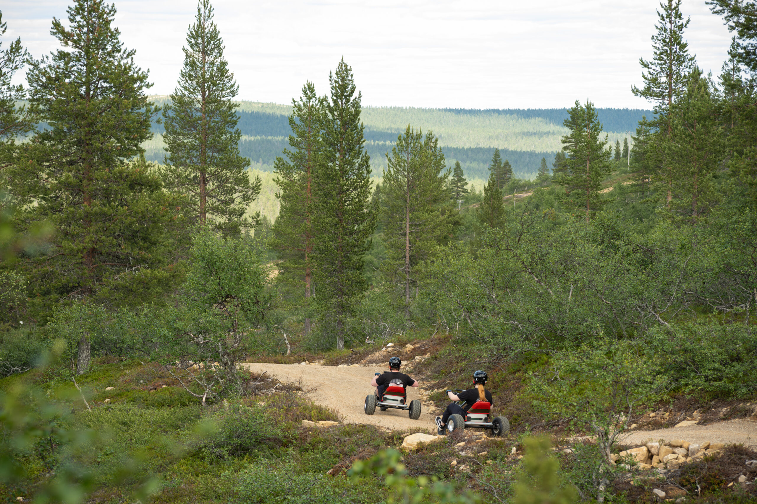 Kaksi ihmistä ajaa polkuautoilla mutkittelevaa hiekkatietä pitkin rehevän vihreän metsän läpi, puiden ja kukkuloiden ympäröimänä pilvisen taivaan alla.