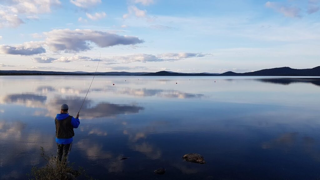 Sinistä takkia pukeutunut henkilö kalastaa rauhallisen järven rannalla; taivaalla on pilviä, ja kaukana olevat kukkulat heijastuvat vedessä sinisen taivaan alla.