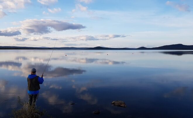 Sinistä takkia pukeutunut henkilö kalastaa rauhallisen järven rannalla; taivaalla on pilviä, ja kaukana olevat kukkulat heijastuvat vedessä sinisen taivaan alla.