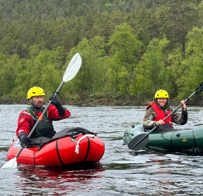 Kaksi kypärää ja pelastusliivejä käyttävää henkilöä meloo ilmatäytteisillä kajakkeilla rauhallisella joella, jota ympäröi tiheä vihreä metsä.