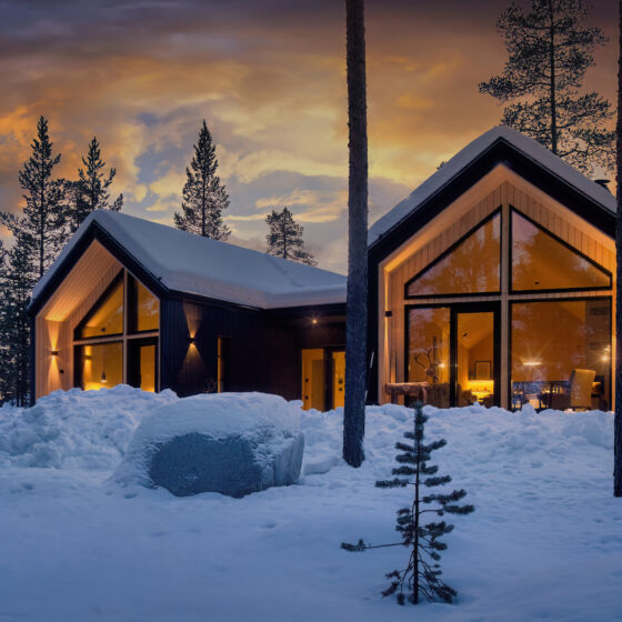 Moderni mökki, jossa on suuret ikkunat, hehkuu lämpimästi auringonlaskun aikaan lumisen metsän keskellä, lumipeitteisten puiden ja rauhallisen talvimaiseman ympäröimänä.