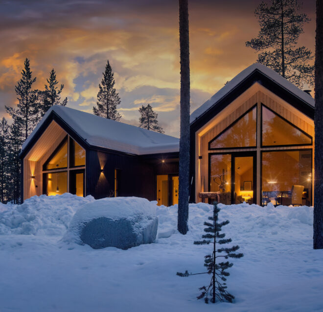 Moderni mökki, jossa on suuret ikkunat, hehkuu lämpimästi auringonlaskun aikaan lumisen metsän keskellä, lumipeitteisten puiden ja rauhallisen talvimaiseman ympäröimänä.