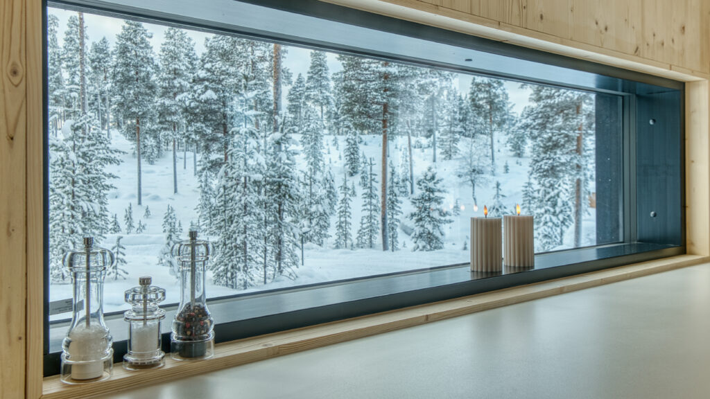 Suuri ikkuna, jonka ikkunalaudalla on kynttilöitä ja maustepurkkeja, avautuu lumiseen metsään, jossa kohoavat korkeat männyt, luoden kodikkaan ja rauhallisen talvisen tunnelman.