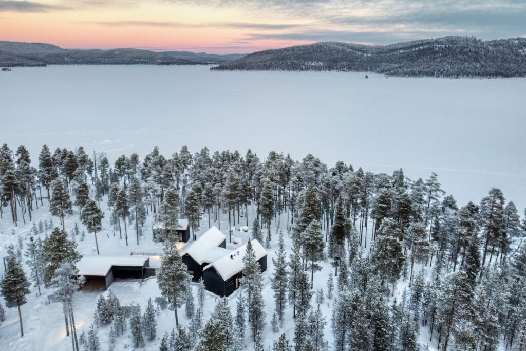 Jäätneen järven rannalla sijaitsee moderni musta talo, jota ympäröivät lumipeitteiset männyt, ja taustalla näkyvät kaukaiset kukkulat sekä pastellisävyinen auringonlaskun taivas.