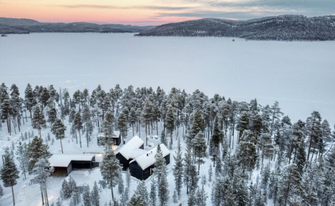 Jäätneen järven rannalla sijaitsee moderni musta talo, jota ympäröivät lumipeitteiset männyt, ja taustalla näkyvät kaukaiset kukkulat sekä pastellisävyinen auringonlaskun taivas.