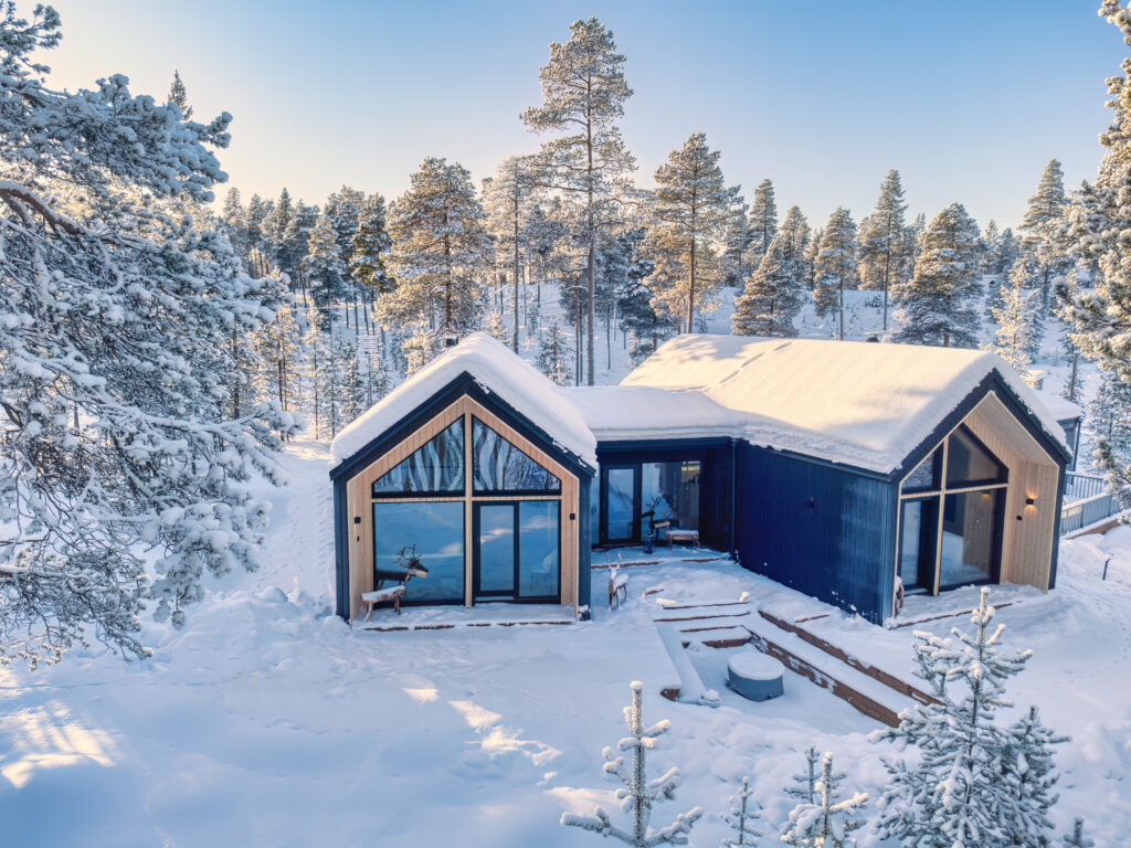 Suurilla ikkunoilla varustettu moderni mökki sijaitsee lumipeitteisten puiden ympäröimänä kirkkaan sinisen taivaan alla, luoden rauhallisen talvimaiseman.