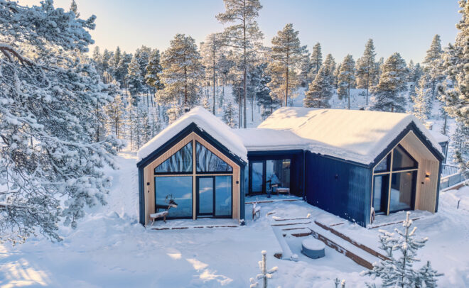 Suurilla ikkunoilla varustettu moderni mökki sijaitsee lumipeitteisten puiden ympäröimänä kirkkaan sinisen taivaan alla, luoden rauhallisen talvimaiseman.