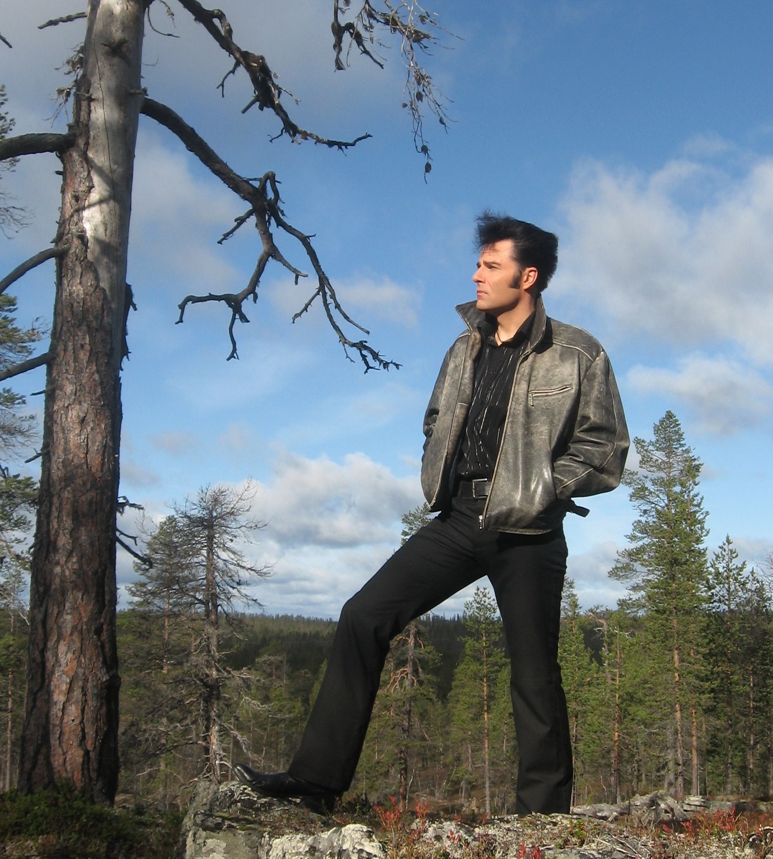 Nahkatakkia ja mustia housuja pukeutunut mies seisoo metsän kalliolla ja katselee kaukaisuuteen kädet taskussaan sinisen taivaan alla, jossa on hajanaisia pilviä.