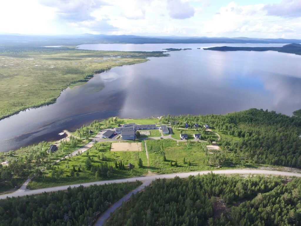 Ilmakuva suuresta järvestä, jota ympäröivät vihreät metsät ja nurmialueet; maisemassa on hajallaan olevia rakennuksia ja sen läpi kulkee tie osittain pilvisen taivaan alla.