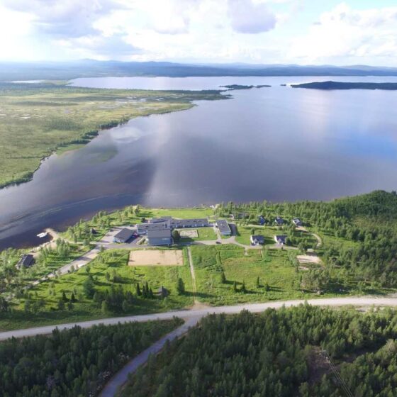 Ilmakuva suuresta järvestä, jota ympäröivät vihreät metsät ja nurmialueet; maisemassa on hajallaan olevia rakennuksia ja sen läpi kulkee tie osittain pilvisen taivaan alla.