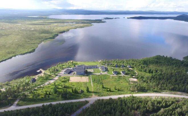 Ilmakuva suuresta järvestä, jota ympäröivät vihreät metsät ja nurmialueet; maisemassa on hajallaan olevia rakennuksia ja sen läpi kulkee tie osittain pilvisen taivaan alla.