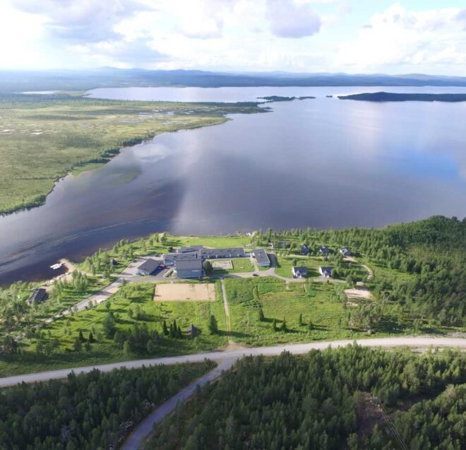 Ilmakuva suuresta järvestä, jota ympäröivät vihreät metsät ja nurmialueet; maisemassa on hajallaan olevia rakennuksia ja sen läpi kulkee tie osittain pilvisen taivaan alla.