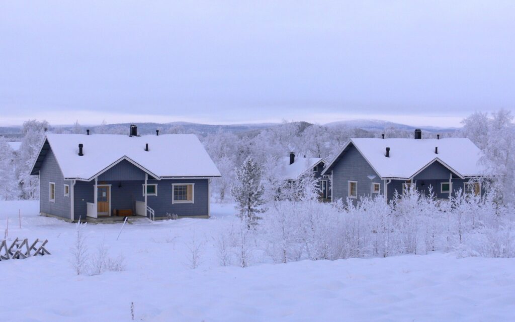 Kaksi sinistä puutaloa, joita ympäröivät lumipeitteiset puut ja maa talvisessa maisemassa; taustalla näkyy kukkuloita pilvisen taivaan alla.