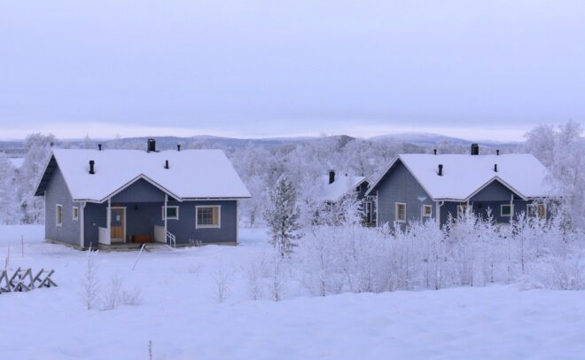 Kaksi sinistä puutaloa, joita ympäröivät lumipeitteiset puut ja maa talvisessa maisemassa; taustalla näkyy kukkuloita pilvisen taivaan alla.