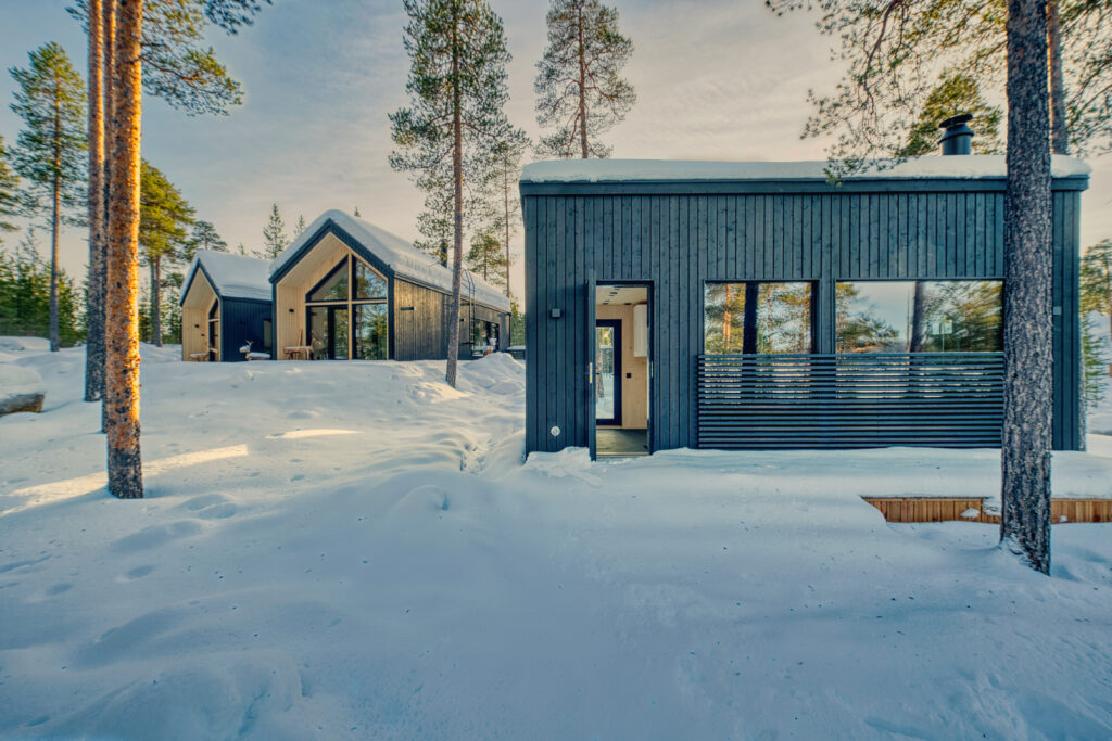 Kaksi modernia mustaa puumökkiä, joissa on suuret ikkunat, sijaitsevat korkeiden mäntyjen keskellä lumisessa maisemassa kirkkaan taivaan alla ja luovat rauhallisen talvimaiseman.