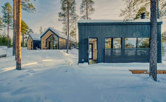Kaksi modernia mustaa puumökkiä, joissa on suuret ikkunat, sijaitsevat korkeiden mäntyjen keskellä lumisessa maisemassa kirkkaan taivaan alla ja luovat rauhallisen talvimaiseman.