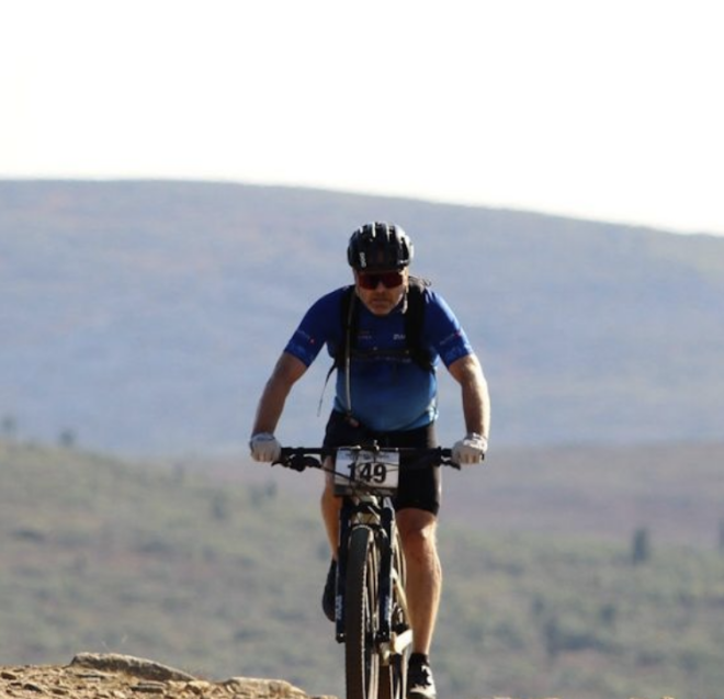 A cyclist wearing a helmet and sunglasses rides a mountain bike on a dirt trail, with hills and sparse trees in the background under a clear sky.