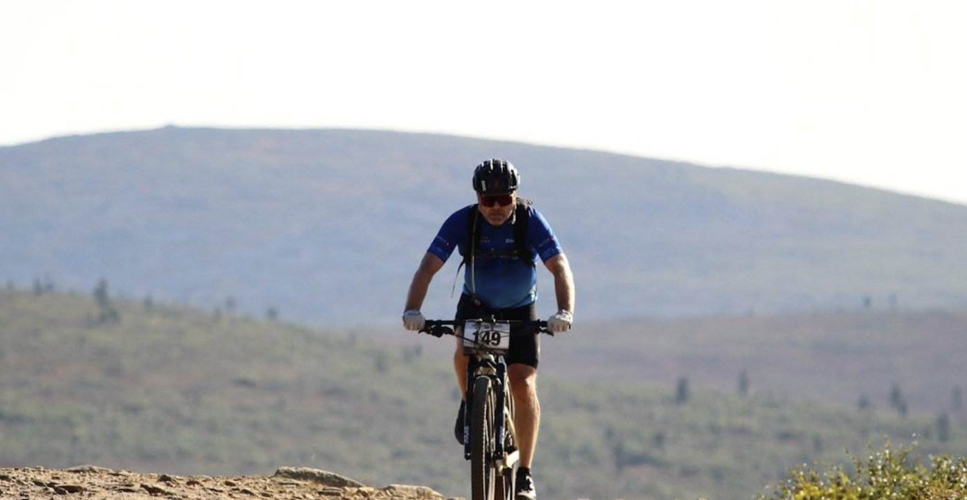 A cyclist wearing a helmet and sunglasses rides a mountain bike on a dirt trail, with hills and sparse trees in the background under a clear sky.