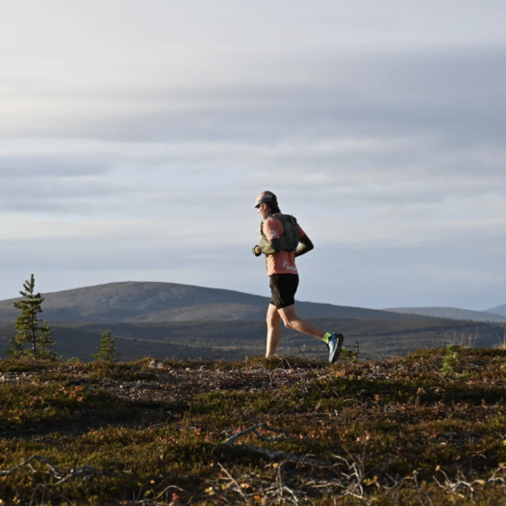 A person in athletic gear runs on a grassy trail with mountains and cloudy skies in the background.
