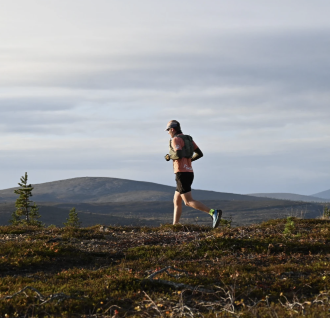 A person in athletic gear runs on a grassy trail with mountains and cloudy skies in the background.