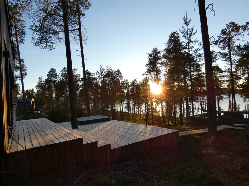 Korkeiden puiden ympäröimältä puuterassilta avautuu näkymä järvelle auringonlaskun aikaan, kun auringonvalo suodattuu puiden läpi ja heijastuu veden pinnalta.
