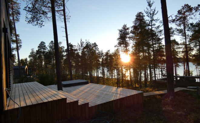 Korkeiden puiden ympäröimältä puuterassilta avautuu näkymä järvelle auringonlaskun aikaan, kun auringonvalo suodattuu puiden läpi ja heijastuu veden pinnalta.