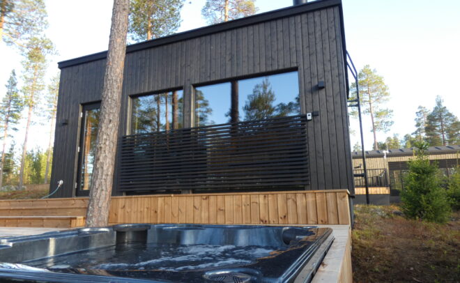 Moderni musta puumökki, jossa on suuret ikkunat ja jota ympäröivät männyt. Etualalla on ulkoilma-kylpytynnyri, jossa on kirkasta vettä ja joka on osittain puuterassin ympäröimä.