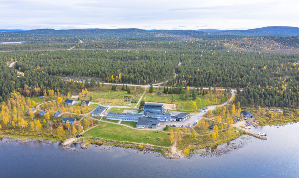 Ilmakuva järvenrannalla sijaitsevasta rakennuskokonaisuudesta, jossa on useita rakennuksia, joita ympäröi metsämaasto ja mutkittelevat tiet, pilvisen taivaan alla. Puiden lehdet ovat syksyn vihreitä ja keltaisia.