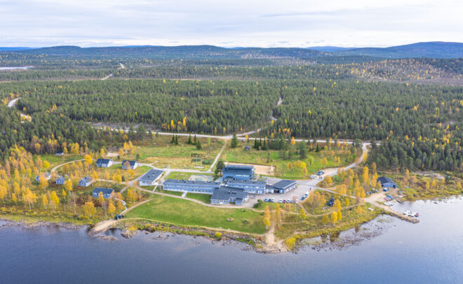Ilmakuva järvenrannalla sijaitsevasta rakennuskokonaisuudesta, jossa on useita rakennuksia, joita ympäröi metsämaasto ja mutkittelevat tiet, pilvisen taivaan alla. Puiden lehdet ovat syksyn vihreitä ja keltaisia.
