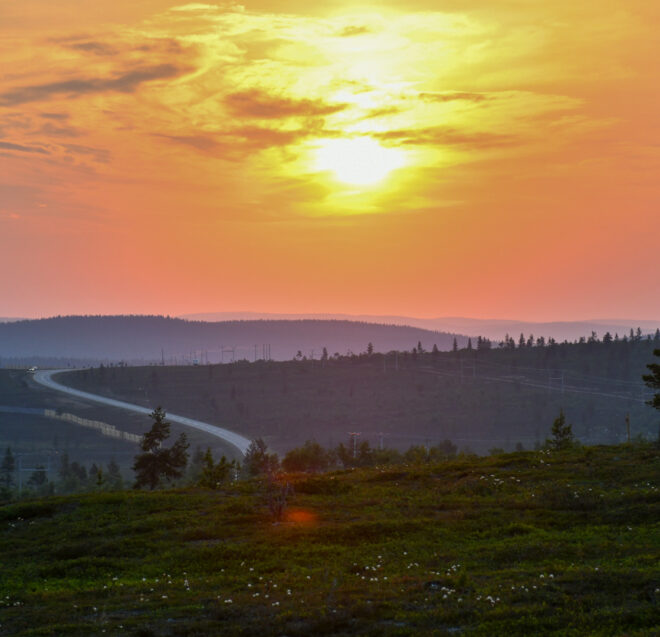 Kiemurteleva tie kulkee mäkisen maiseman läpi auringonlaskun aikaan; taivas on oranssi ja keltainen, pilviä on hajallaan ja etualalla näkyy puiden siluetteja.