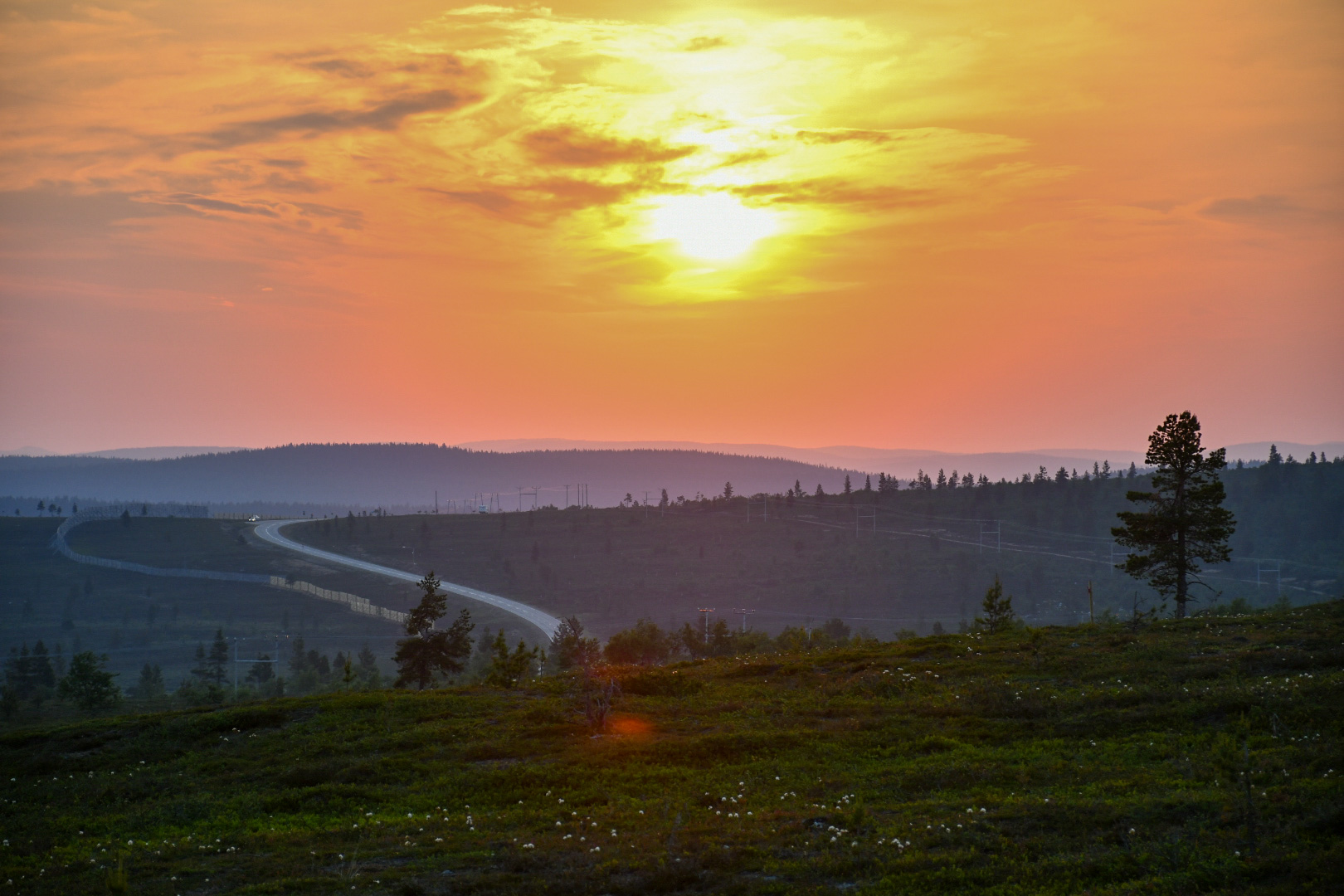 Kiemurteleva tie kulkee mäkisen maiseman läpi auringonlaskun aikaan, kun taivas on oranssi ja pilviä on hajallaan. Puiden siluetit näkyvät etualalla ja kaukaisilla kukkuloilla.