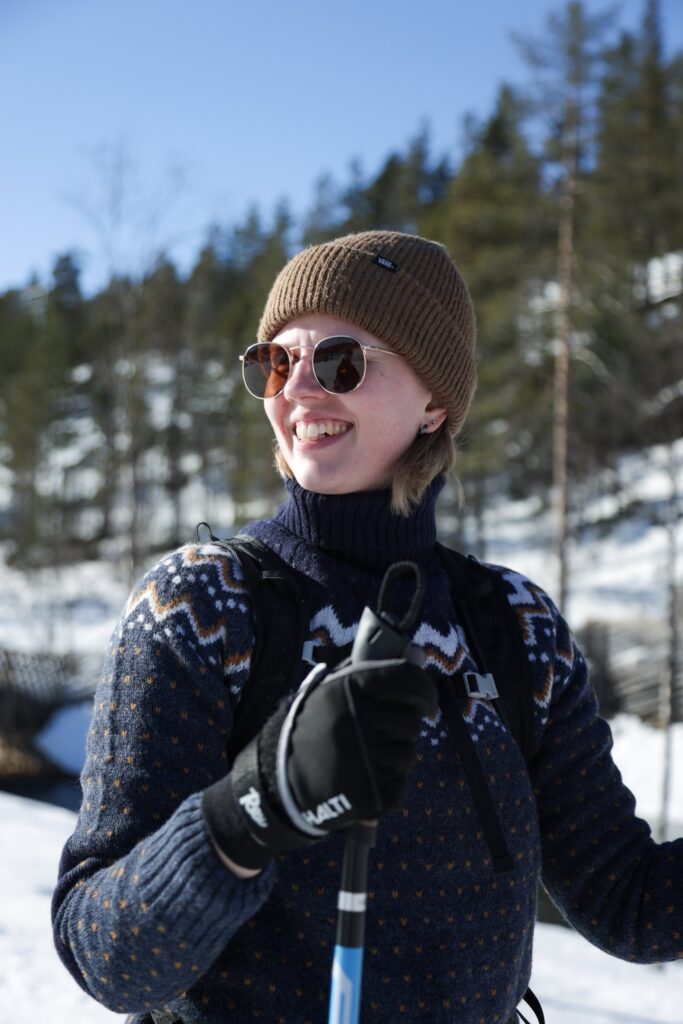 Eine Person mit Sonnenbrille, brauner Mütze, Handschuhen und einem gemusterten Pullover lächelt, während sie im Freien in einer verschneiten, sonnigen Loipenlandschaft in Lappland mit Bäumen im Hintergrund ihre Skistöcke in den Händen hält.